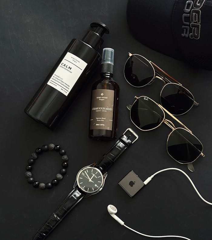 Flat lay of men's accessories including sunglasses, watch, and skincare products on black background.