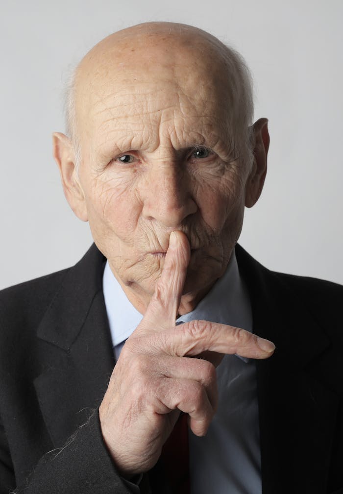 Portrait of a senior man wearing a suit, gesturing for silence in a studio setting.